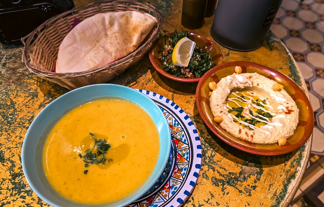A bowl of yellow soup, hummus, and pita bread on a rustic table